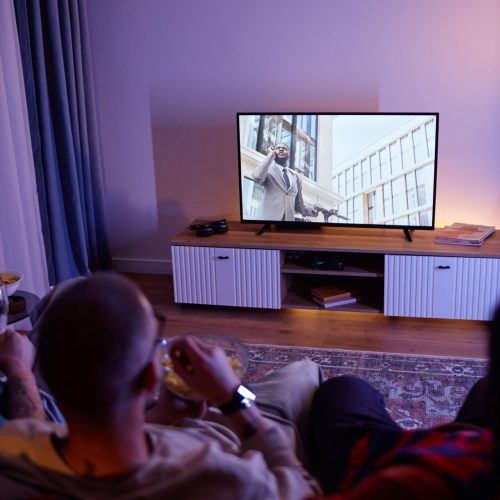 Vertical back view of friends watching Tv at night in dark room while sitting on sofa with focus on TV screen, copy space